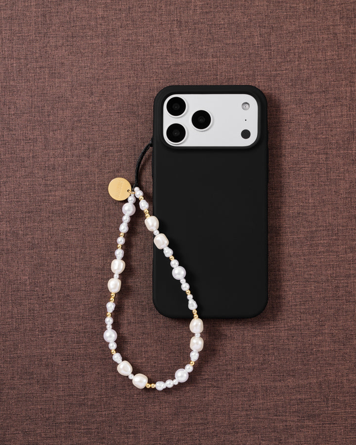 White baroque faux-pearl and gold-bead phone strap looped on a black phone case, styled flat on mauve textured fabric.