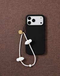 White cloud beaded phone strap looped beside a black phone case on woven brown fabric, gold-tone disc and cloud beads on show.