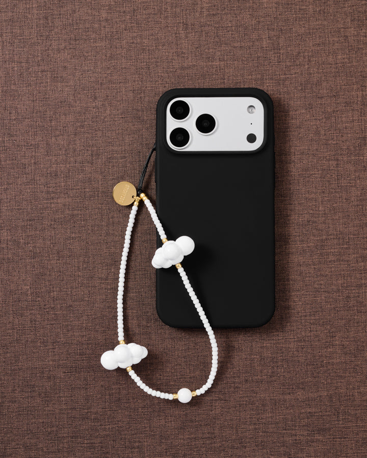 White cloud beaded phone strap looped beside a black phone case on woven brown fabric, gold-tone disc and cloud beads on show.