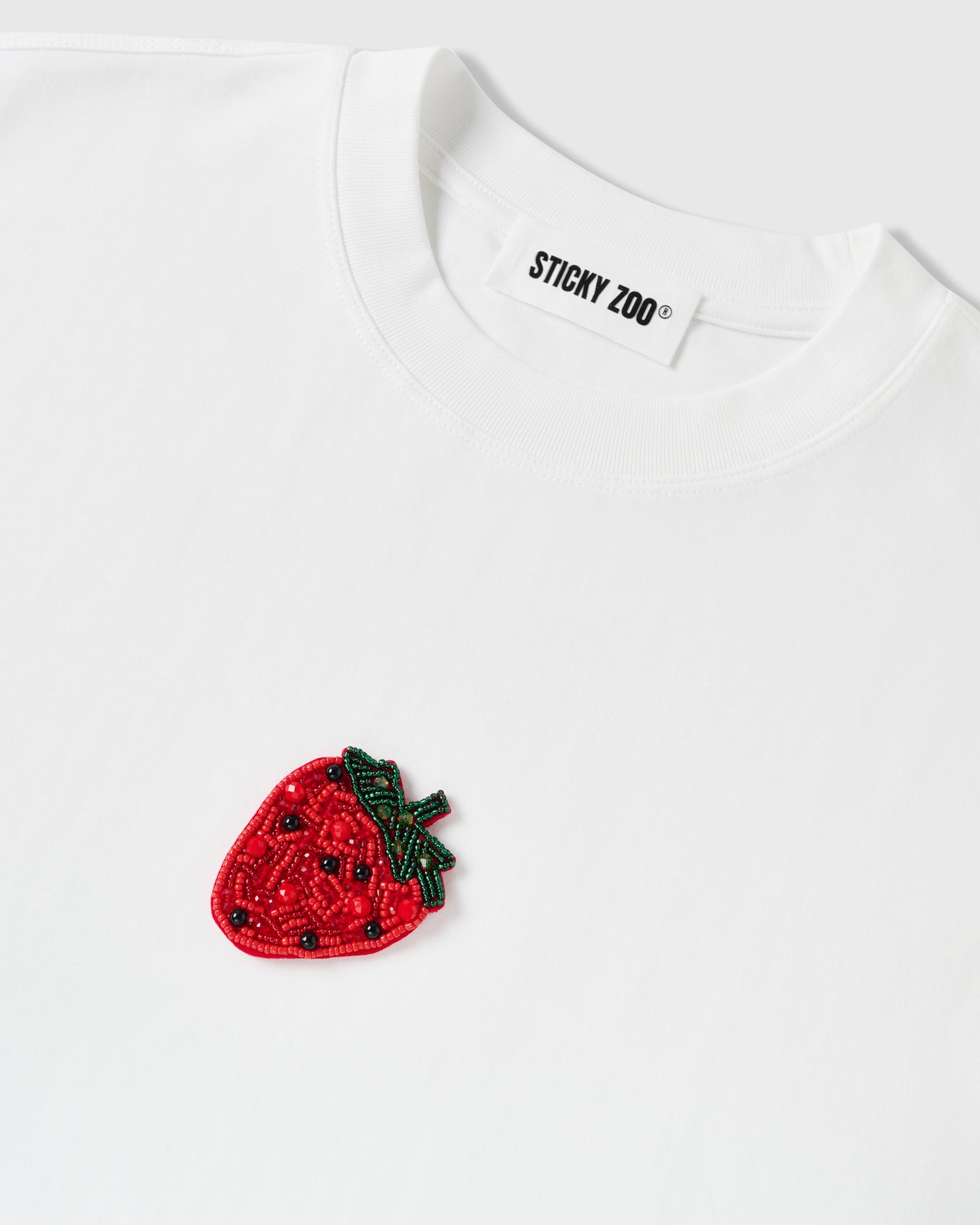Macro of dense hand-sewn beadwork on the STICKYZOO Strawberry T-Shirt with the woven neck label, showing hundreds of individual glass beads