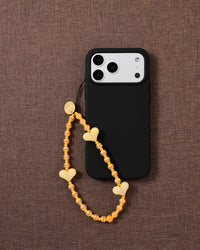 Yellow heart beaded strap looped onto a black phone case on brown fabric, hearts and orange-striped beads draping from a gold-tone disc.