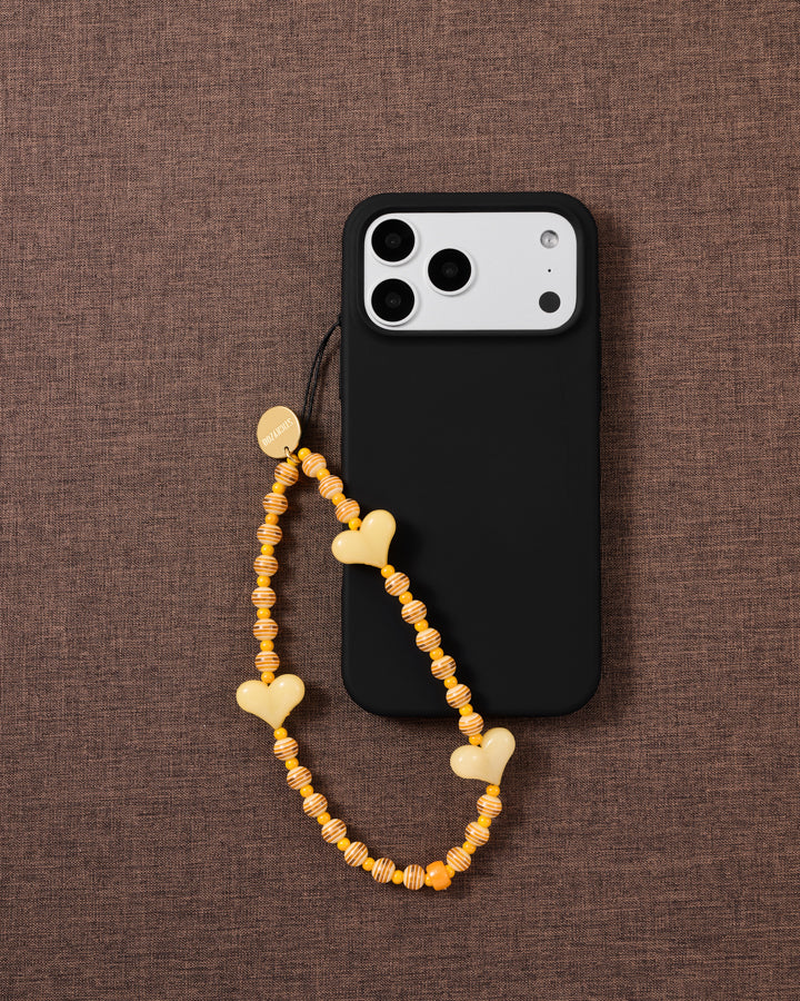 Yellow heart beaded strap looped onto a black phone case on brown fabric, hearts and orange-striped beads draping from a gold-tone disc.