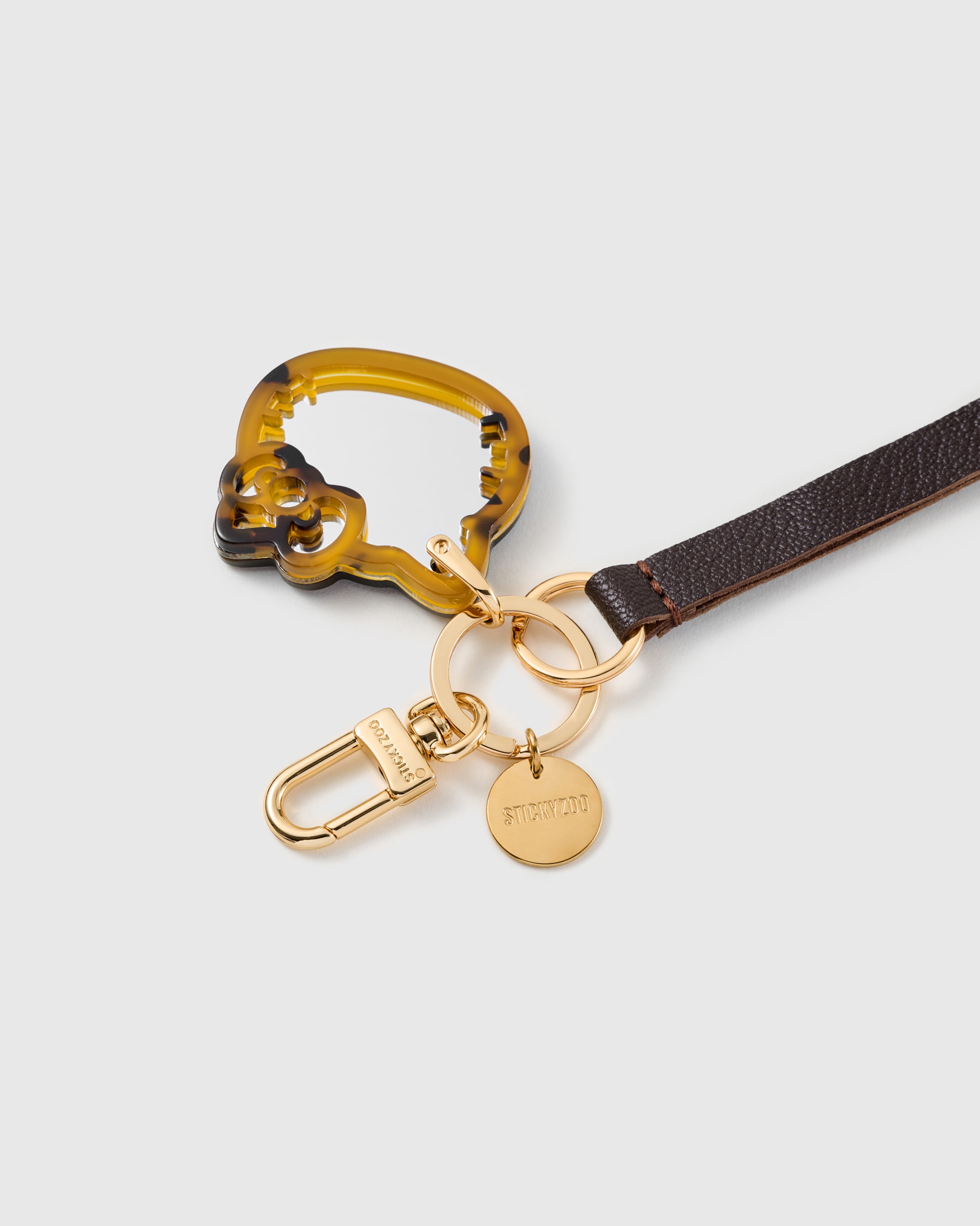 Close-up of tortoise acetate cat mirror charm showing layered amber and brown pattern with engraved STICKYZOO gold hardware