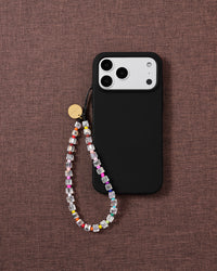 Clear beaded phone strap looped onto a black phone case on textured mauve fabric, rainbow seed beads and a gold disc catching the light.
