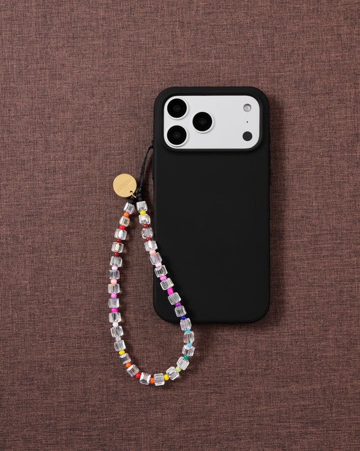 Clear beaded phone strap looped onto a black phone case on textured mauve fabric, rainbow seed beads and a gold disc catching the light.