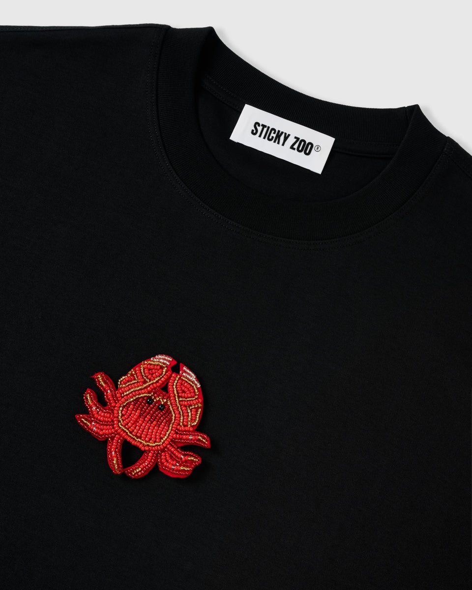 Detail of the beaded crab applique beside the ribbed crew neck and STICKYZOO neck label, showing raised glass-bead texture.