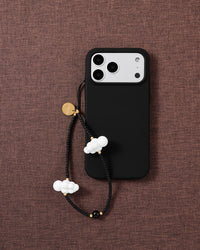 Cloud beaded phone strap looped on a black phone case over brown textile, showing two white clouds, matte black beads and a gold disc.