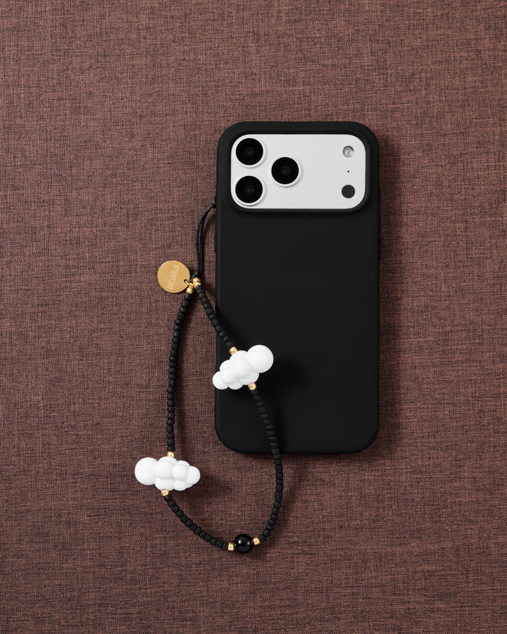 Cloud beaded phone strap looped on a black phone case over brown textile, showing two white clouds, matte black beads and a gold disc.