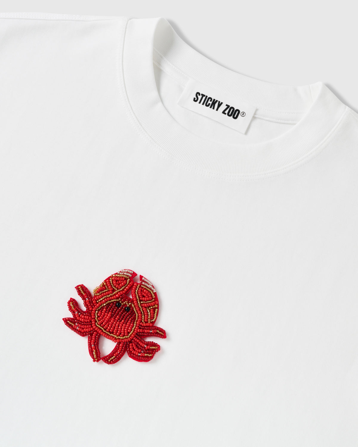 Detail of the beaded crab applique beside the ribbed crew neck and STICKYZOO neck label on the white tee, showing raised glass-bead texture.