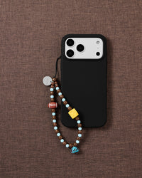 Beaded loop strap looped onto a black phone case over brown linen, showing the football, cheese bead and blue puppy bell on cocoa and sky-blue beads.