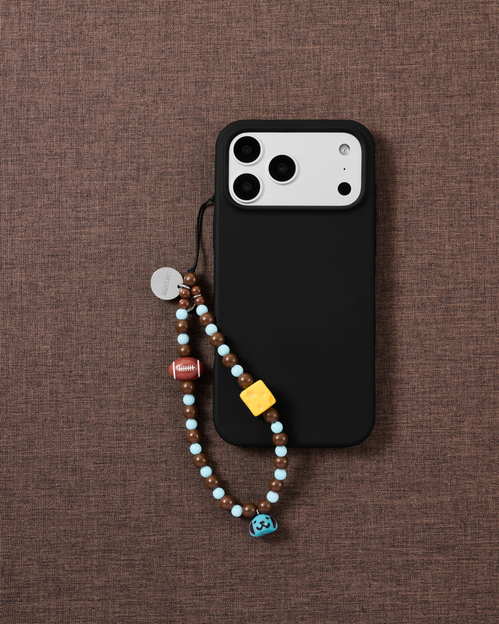 Beaded loop strap looped onto a black phone case over brown linen, showing the football, cheese bead and blue puppy bell on cocoa and sky-blue beads.