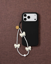 The ivory bow strap looped on a black phone case, faux-pearl beads and cream bows resting on mauve fabric