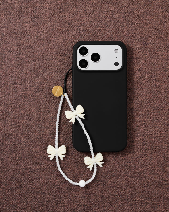 The ivory bow strap looped on a black phone case, faux-pearl beads and cream bows resting on mauve fabric