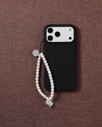 Iridescent glitter bead phone strap with a clear heart drop looped on a black phone case, resting on dusty-rose fabric.