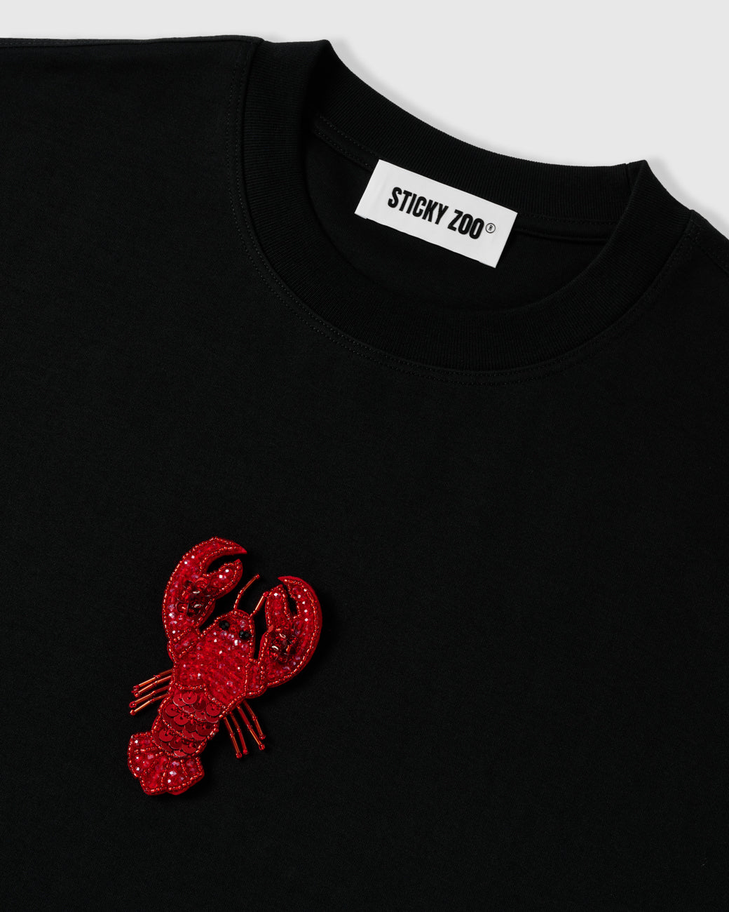 Detail of the beaded lobster applique beside the ribbed crew neck and STICKYZOO neck label, showing raised glass-bead and sequin texture.