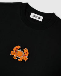 Angled close-up of the STICKYZOO beaded crab appliqué and ribbed crew neck showing raised seed-bead texture on matte black cotton