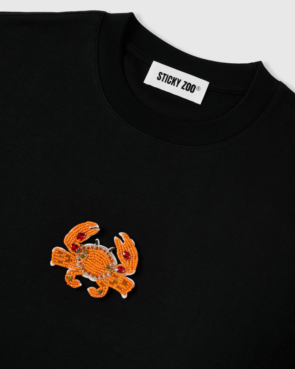 Angled close-up of the STICKYZOO beaded crab appliqué and ribbed crew neck showing raised seed-bead texture on matte black cotton