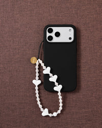 STICKYZOO Snow Heart beaded strap looped on a black phone case over brown fabric, white hearts and ribbed cream-white discs with a gold-tone disc.
