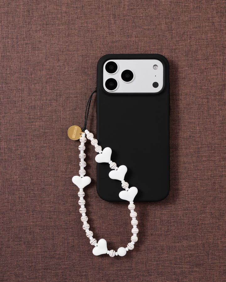 STICKYZOO Snow Heart beaded strap looped on a black phone case over brown fabric, white hearts and ribbed cream-white discs with a gold-tone disc.