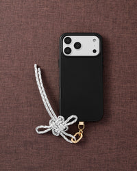 Pearl-white braided bow phone strap with gold hardware on textured brown fabric lifestyle flat-lay
