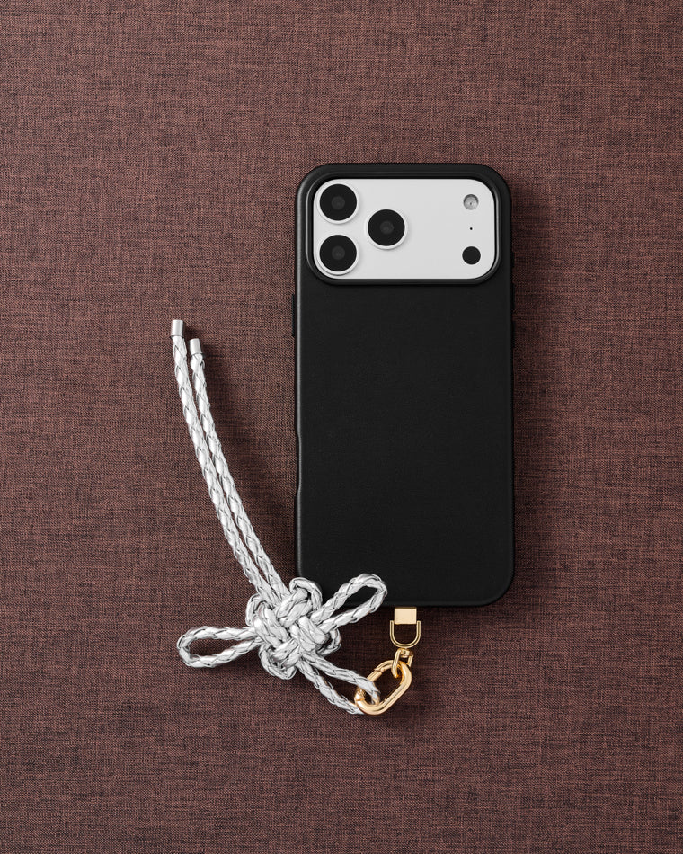 {{Designed in Amsterdam}} Pearl-white braided bow phone strap with gold hardware on textured brown fabric lifestyle flat-lay