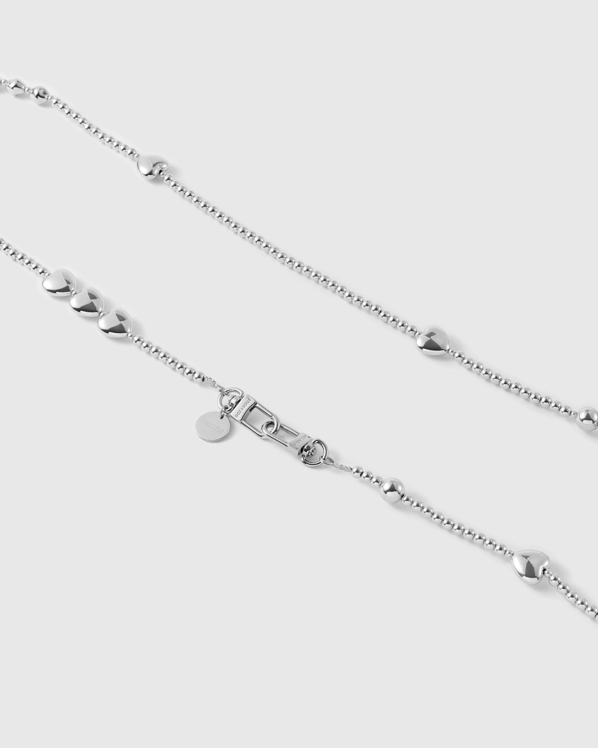 Close-up of the silver-tone ball-bead chain showing a graduated cluster, a puffy heart, and the dual clasp beside a round engraved disc.