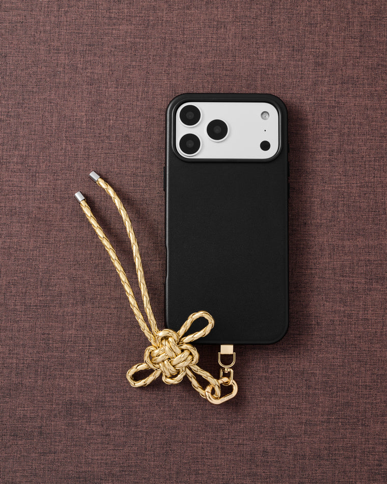 {{Designed in Amsterdam}} Champagne gold bow phone strap on dark brown fabric lifestyle flat-lay with black phone case