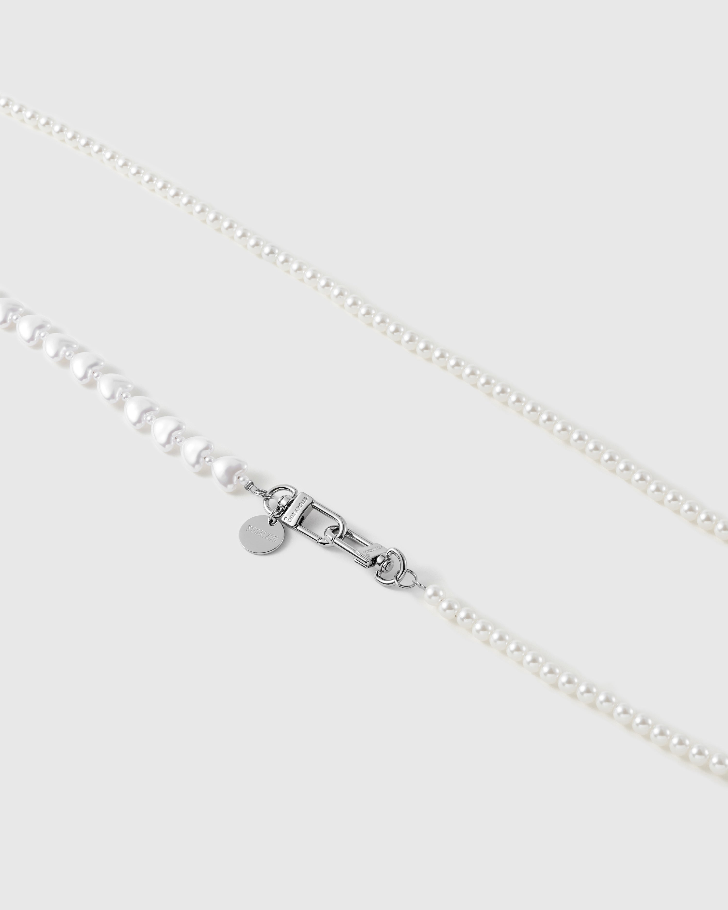 Close-up of round white faux-pearls meeting puffy heart beads beside a silver-tone swivel clasp and a small engraved disc.