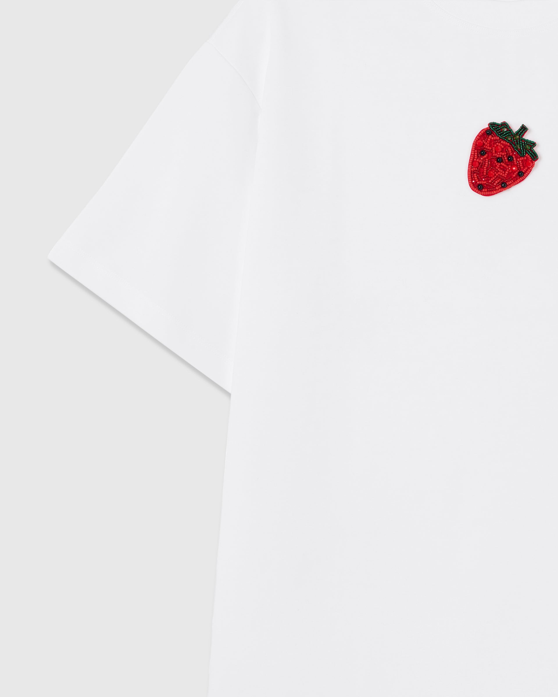 Angled close-up of the hand-beaded strawberry and ribbed crew neck on the white oversized cotton tee, faceted beads catching the light