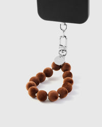 Toffee pom phone strap hanging from black phone case showing wrist carry with silver hardware