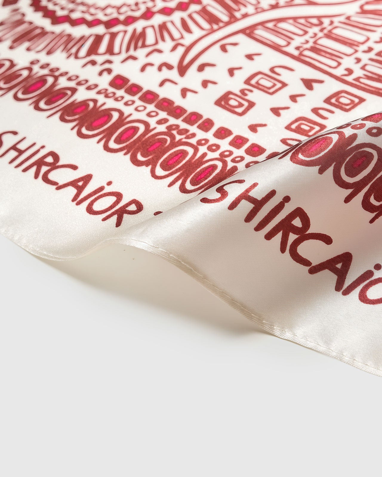 Silk scarf folded to reveal the cream field, crimson line work, and the lettered border catching the light
```