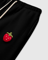 Close-up of hand-stitched 3D plush strawberry at hip showing stuffed texture and green stem on black cotton