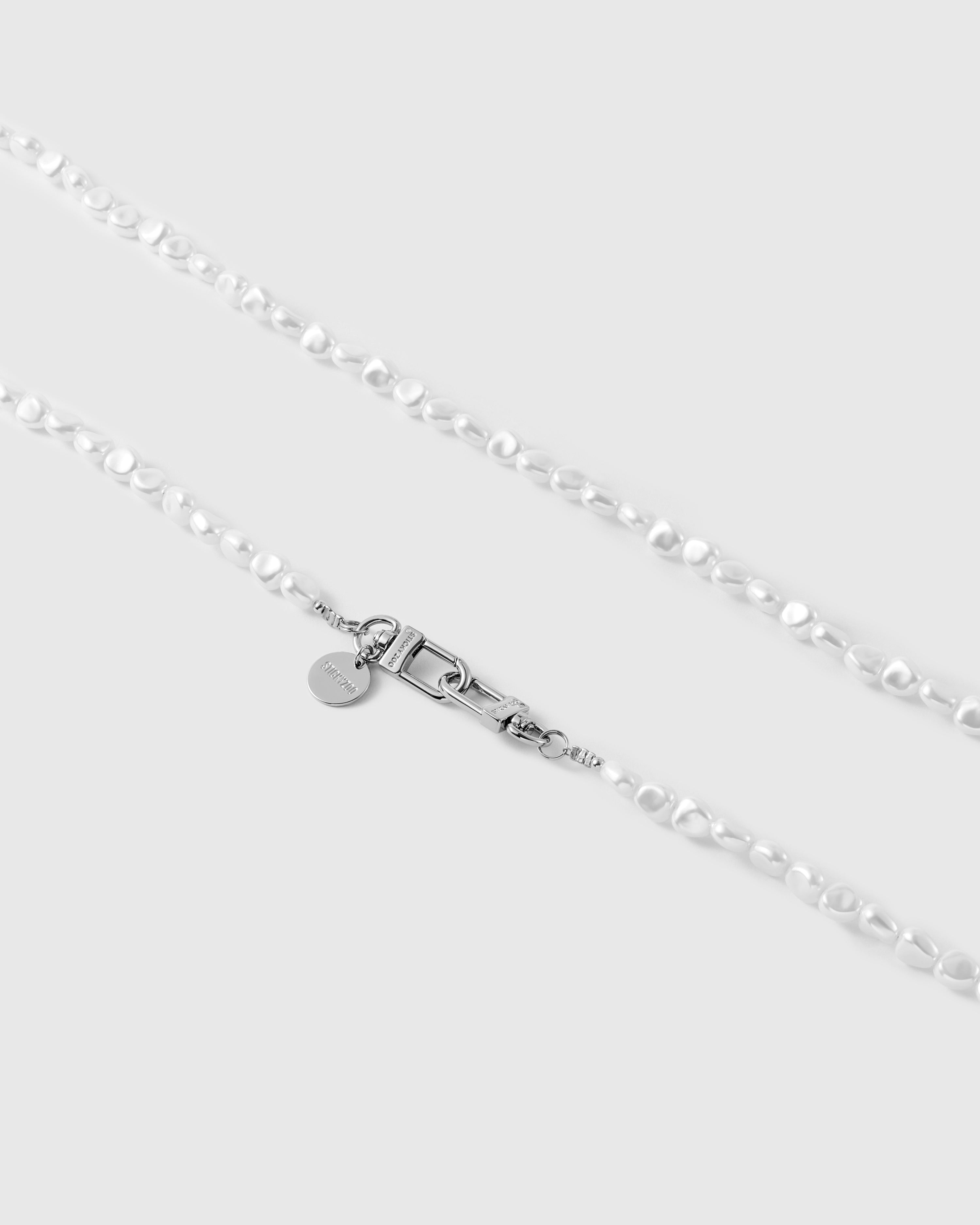 Close-up of silver-tone dual lobster clasps and an engraved round STICKYZOO disc set against irregular white faux-pearl beads.