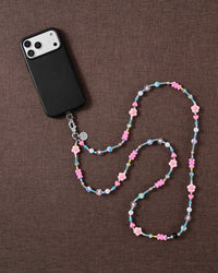 STICKYZOO cross-body phone strap on brown linen, a black phone at one end and pink flower, heart, pearl and turquoise beads curving across.