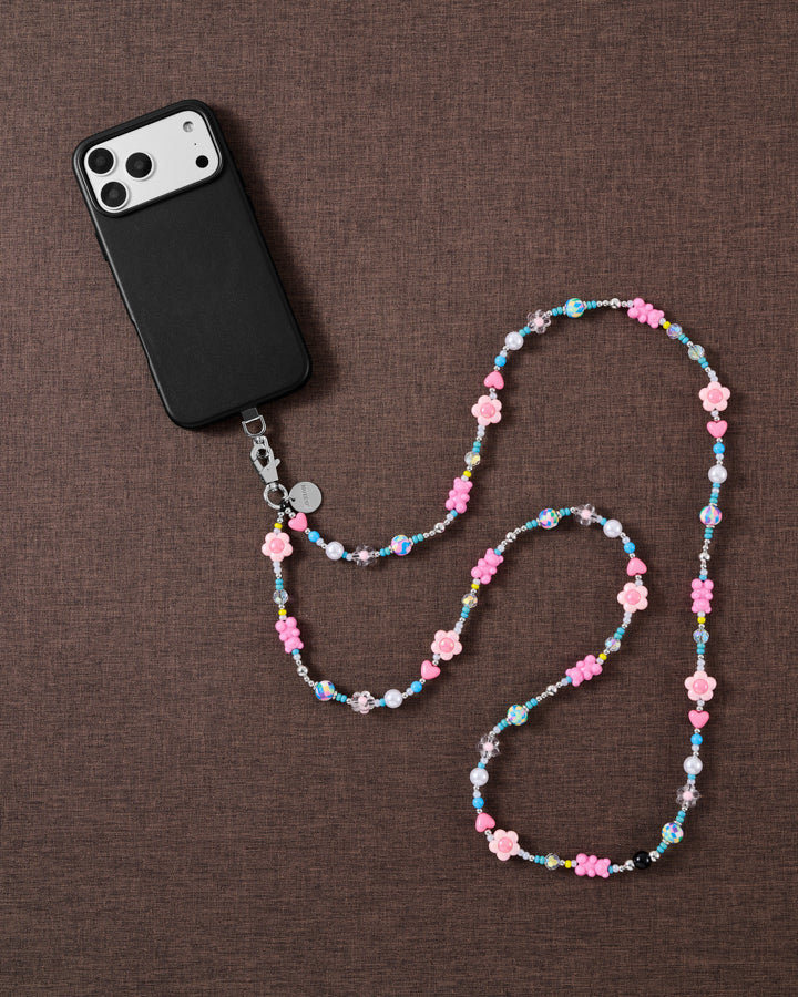 STICKYZOO cross-body phone strap on brown linen, a black phone at one end and pink flower, heart, pearl and turquoise beads curving across.