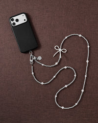 Flat-lay of the STICKYZOO beaded cross-body strap on brown linen beside a black phone, silver-tone beads, faux-pearls and a faux-pearl bow.