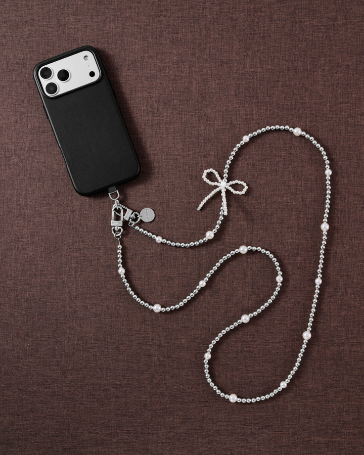 Flat-lay of the STICKYZOO beaded cross-body strap on brown linen beside a black phone, silver-tone beads, faux-pearls and a faux-pearl bow.
