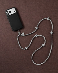 Silver-tone puffy-heart bead crossbody strap arranged beside a black phone case on brown textured fabric.