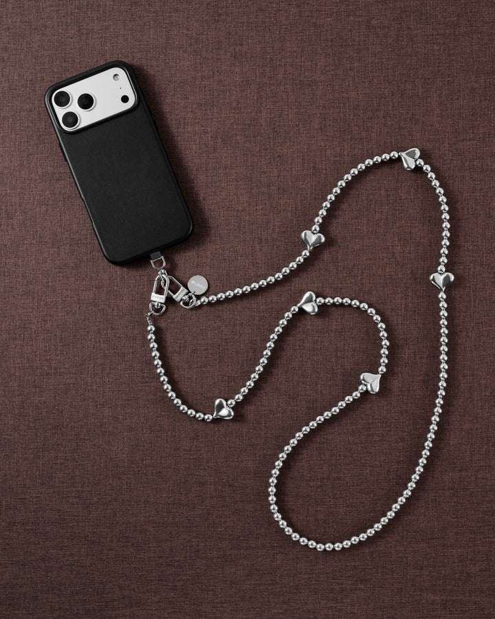 Silver-tone puffy-heart bead crossbody strap arranged beside a black phone case on brown textured fabric.