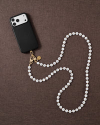 Flat-lay on brown linen: black phone case and the full white faux-pearl cross-body strand curving in an S, gold clasp beside it.