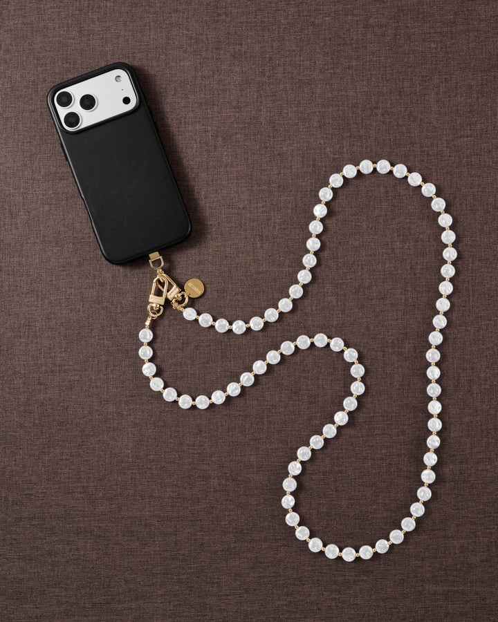 Flat-lay on brown linen: black phone case and the full white faux-pearl cross-body strand curving in an S, gold clasp beside it.