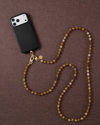 Styled flat-lay of a black phone on brown fabric, the honey-amber STICKYZOO crossbody strap laid in a soft S-curve with gold-tone clasps.