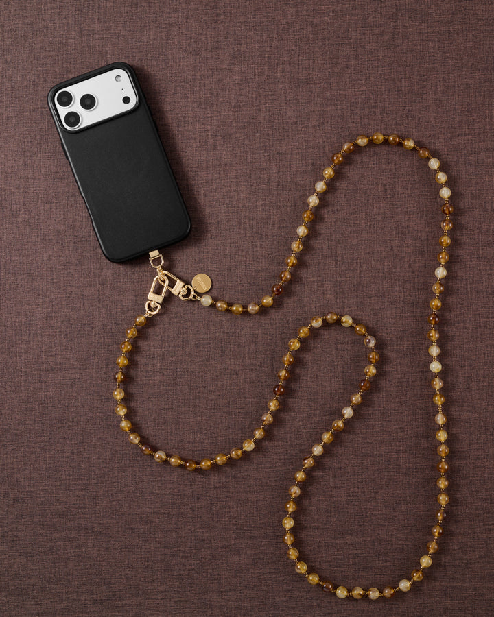 Styled flat-lay of a black phone on brown fabric, the honey-amber STICKYZOO crossbody strap laid in a soft S-curve with gold-tone clasps.