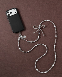STICKYZOO cross-body strap styled flat with a black phone on brown fabric: silver balls, faux-pearls, clear beads and a woven bow.