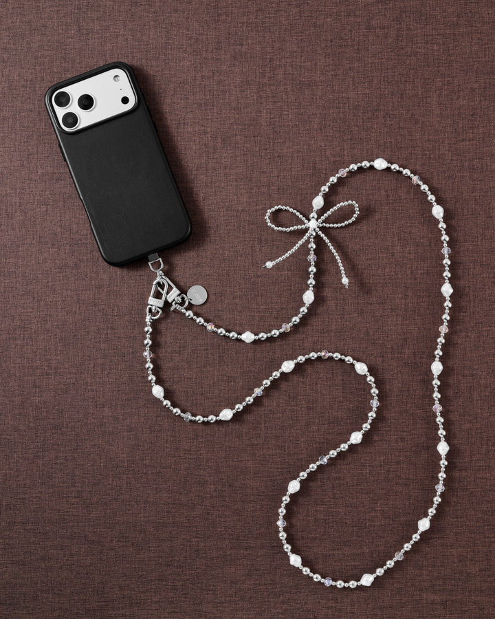 STICKYZOO cross-body strap styled flat with a black phone on brown fabric: silver balls, faux-pearls, clear beads and a woven bow.