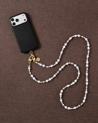 Pearl-and-gold crossbody strap laid in an S-curve on brown linen beside a black phone case, showing its full worn length.