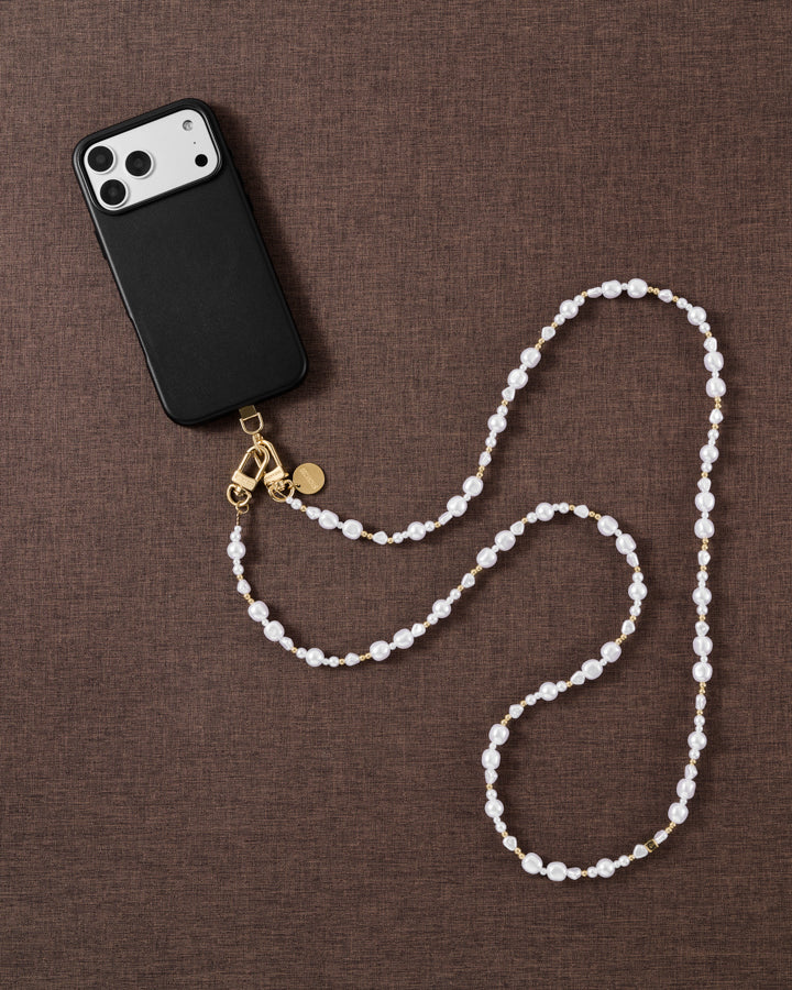 Pearl-and-gold crossbody strap laid in an S-curve on brown linen beside a black phone case, showing its full worn length.