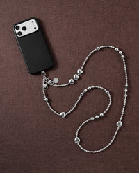 Silver-tone STICKYZOO cross-body strap styled beside a black phone case on brown linen, ball-bead chain curving with hearts and clusters.