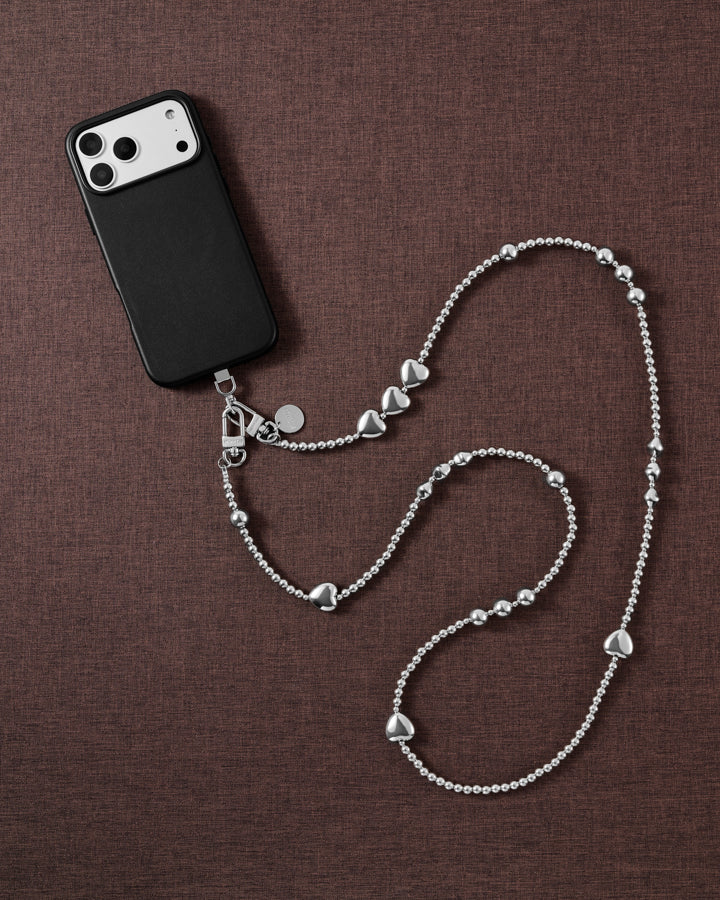 Silver-tone STICKYZOO cross-body strap styled beside a black phone case on brown linen, ball-bead chain curving with hearts and clusters.