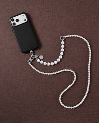 Styled flat-lay on brown fabric: a black phone with the white pearl-and-heart strap draped crossbody-length, silver-tone clasp and disc.